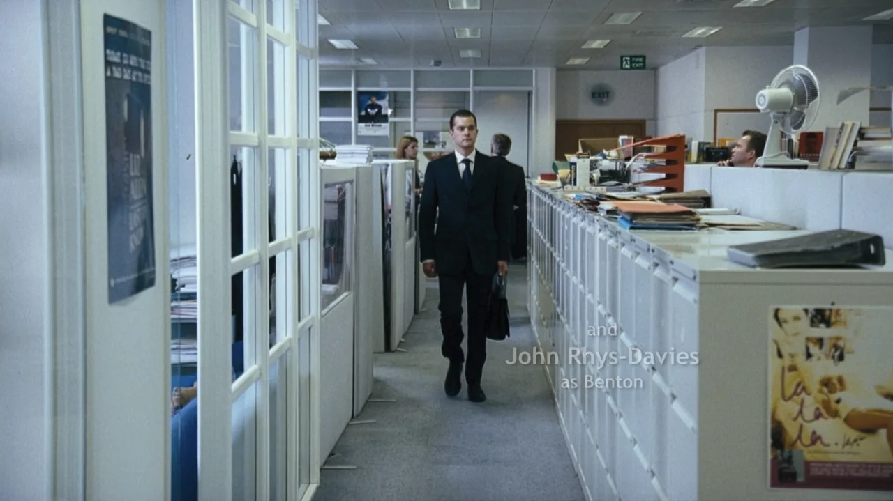 Shadows in the Sun (2005) movie still: fullbody — A corporate workspace features rows of white cubicle dividers under bright overhead lighting. A…