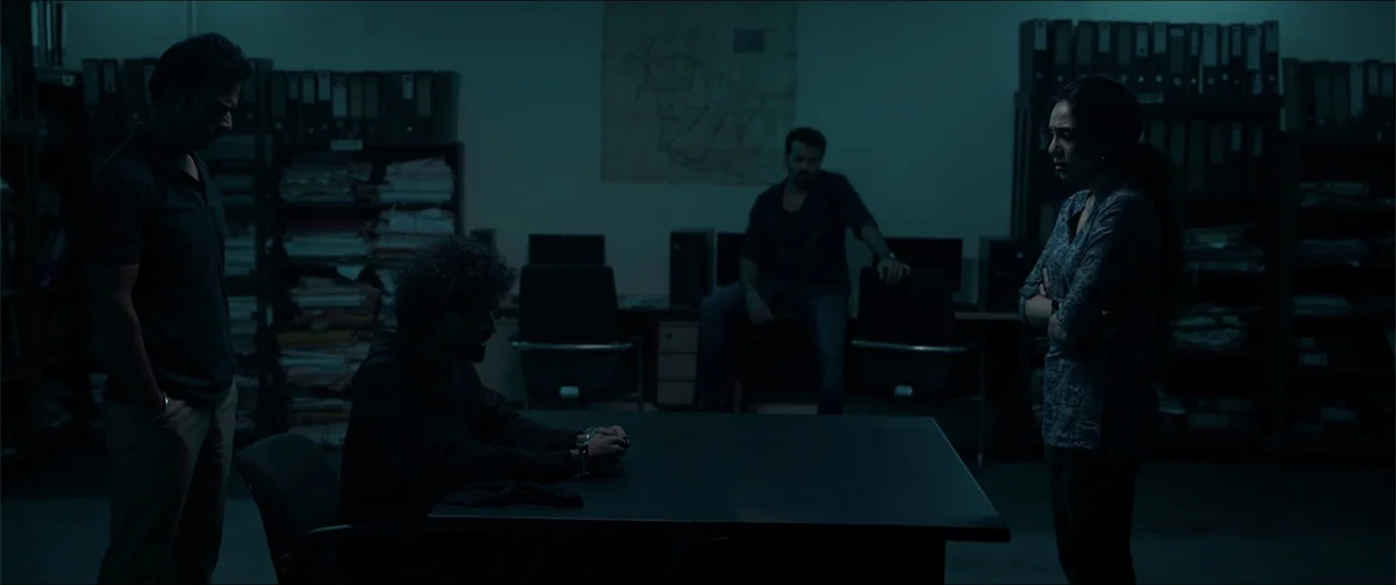Cheekatilo (2026) movie still: wide — A dimly lit room contains rows of file folders on shelves against the back wall. Four people gather in the…