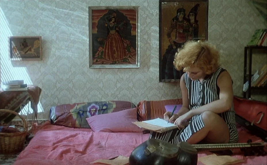 L’une Chante, l’Autre Pas (1977) movie still: medium — A woman sits on a bed covered in a pink blanket against patterned wallpaper. She writes on a…