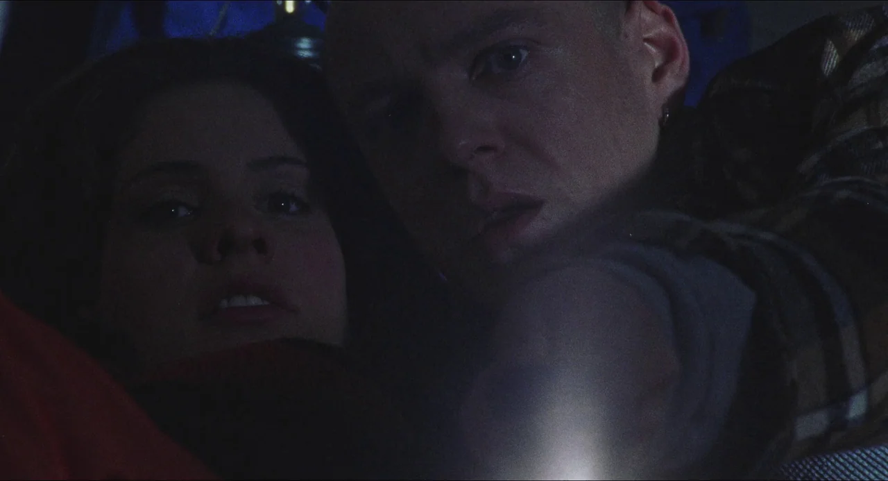 Dog Soldiers (2002) movie still: closeup — A man and woman are closely positioned in a dark, shadow-filled indoor space. They look toward an unseen…