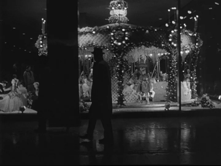 Blast Of Silence (1961) movie still: fullbody — A nighttime city sidewalk passes a large window filled with a glowing carousel display. A man…