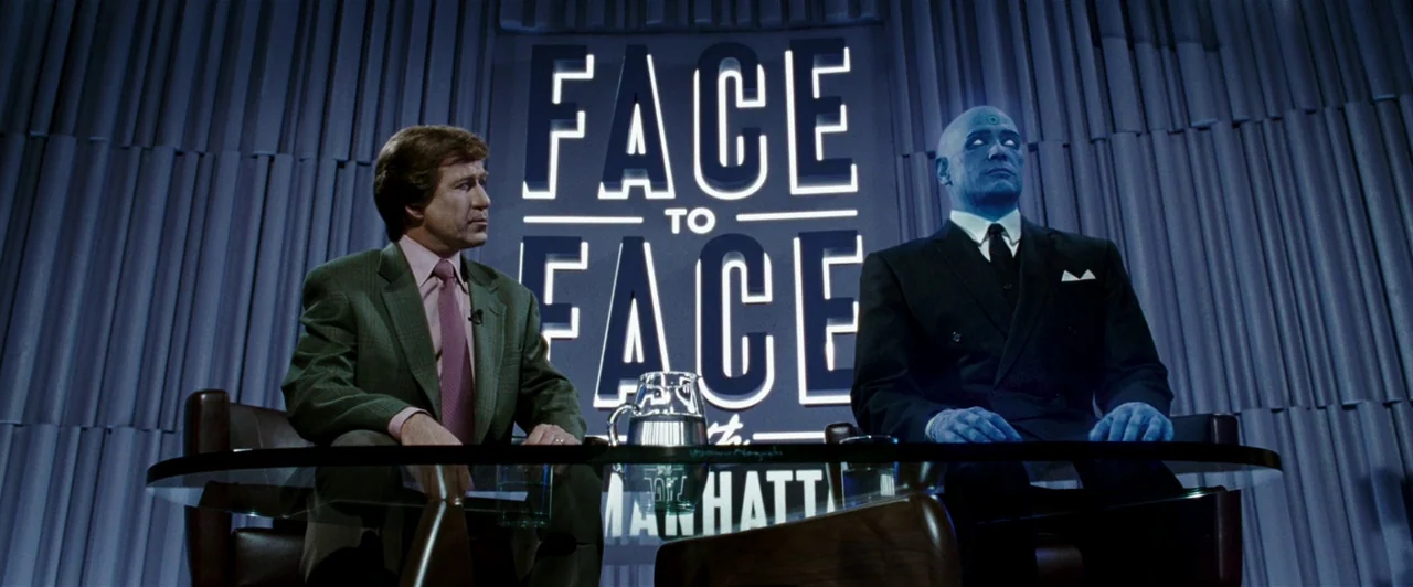 Watchmen (2009) movie still: medium — A television interview set features a desk in front of a vertical paneled background. Two men sit at the desk…