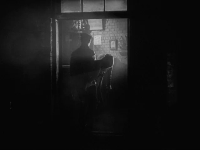 The Docks of New York (1928) movie still: medium — A dark space looks into a brightly lit room featuring a brick wall. A man carries an unconscious…