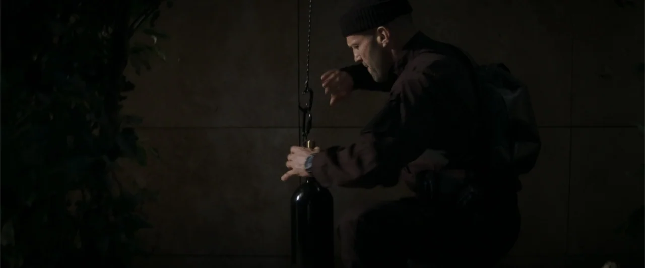 Operation Fortune: Ruse de Guerre (2023) movie still: medium — A dark outdoor space features a concrete wall and a hanging metal canister. A man in…
