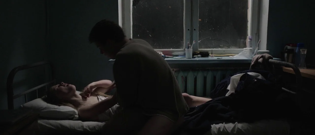 The Tribe (2014) movie still: medium — A dimly lit room at night features a bed placed directly beneath a window. A man sits over a woman while she…