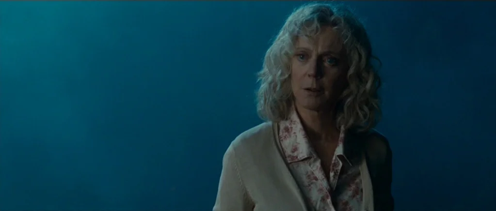 Paul (2011) movie still: medium — A woman stands in a dimly lit space against a flat blue wall. The subject gazes toward the left side of the frame…