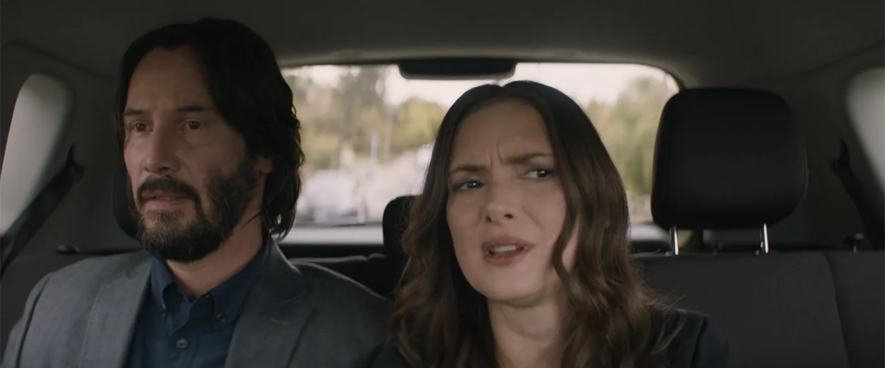 Destination Wedding (2018) movie still: medium — Two individuals sit in the back row of a moving car during daylight hours. A man with a dark beard…