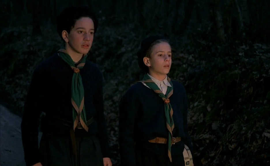 Au Revoir Les Enfants (1987) movie still: medium — Two boys dressed in scout uniforms stand on a dark, tree-lined path at twilight. They gaze…
