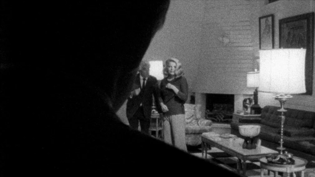 Faces (1968) movie still: medium — A dimly lit room with mid-century furniture and lamps illuminating the space. A man and a woman stand in the…