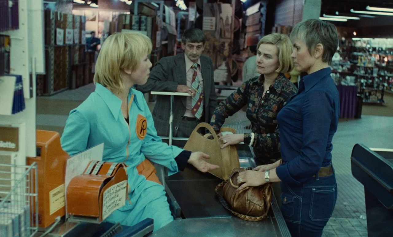 Graduate First (1978) movie still: medium — A retail checkout counter sits inside a large store with aisles of goods. A female cashier interacts…