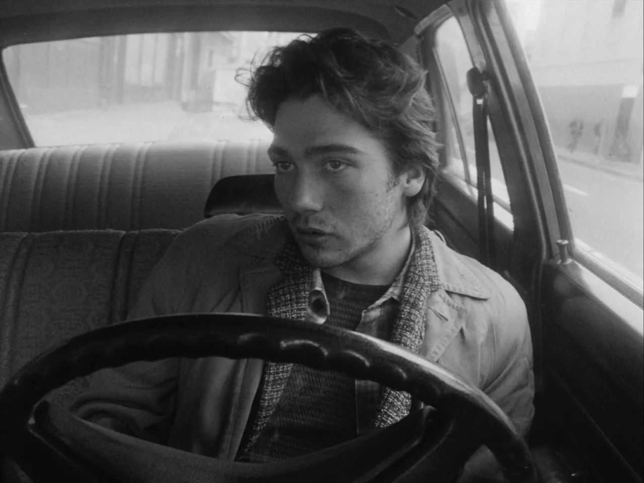 Mala Noche (1986) movie still: medium — A man sits in the driver seat of a vintage car during the day. He grips the steering wheel while looking out…