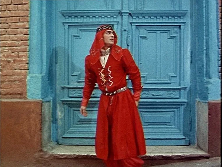 Ashik Kerib (1988) movie still: fullbody — A man in a vibrant red costume stands before a large bright blue door. The structure is flanked by…