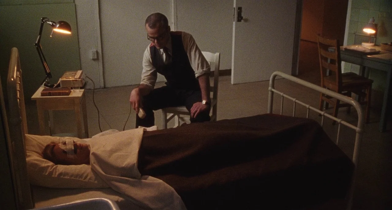 Chained For Life (2018) movie still: medium — A stark hospital room features a metal bed frame and simple wooden furniture. A man in formal work…