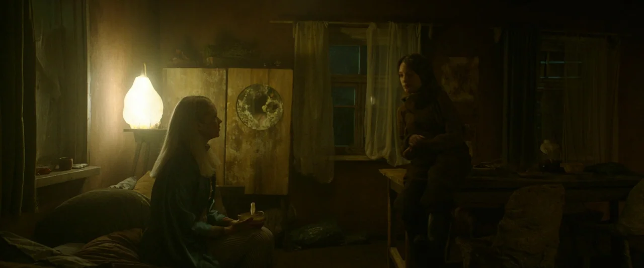 Vesper (2022) movie still: medium — A dimly lit room with worn wooden furniture and thin, tattered window curtains. Two figures sit in a sparse,…
