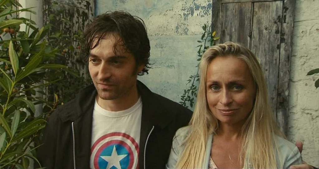 The Beaches of Agnès (2008) movie still: medium — An outdoor area with peeling white paint on a wall and weathered wooden panels. A man and a woman…