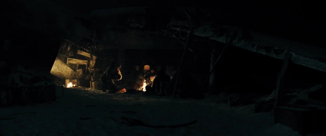 The Grey (2012) movie still: wide — A group of people huddle around a small fire in a dark, snowy landscape. The wreckage of a structure or vehicle…