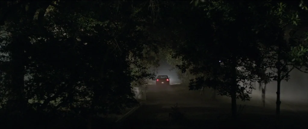 Abattoir (2016) movie still: wide — A dark, dirt-covered road is flanked by dense, silhouetted trees under a heavy night fog. A solitary car drives…