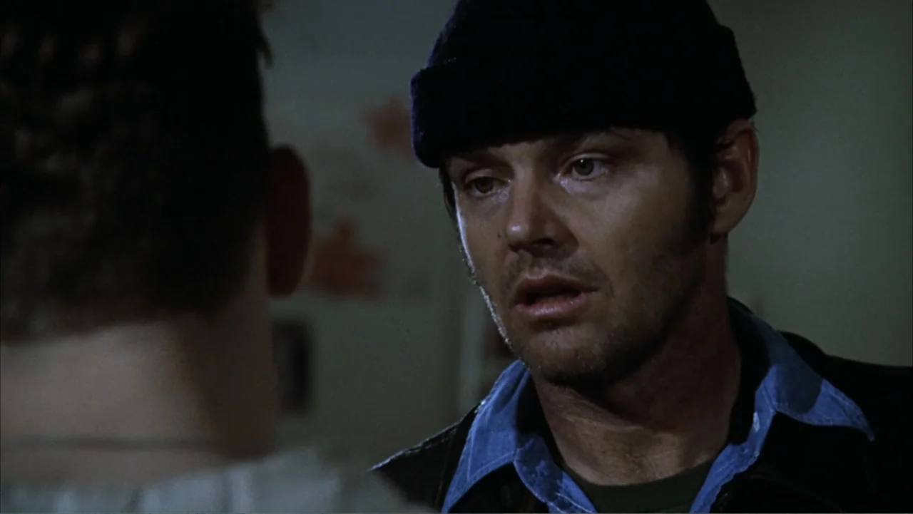 One Flew Over the Cuckoo's Nest (1975) movie still: medium — A dimly lit room with muted tones and a blurred foreground shoulder. A man wearing a…