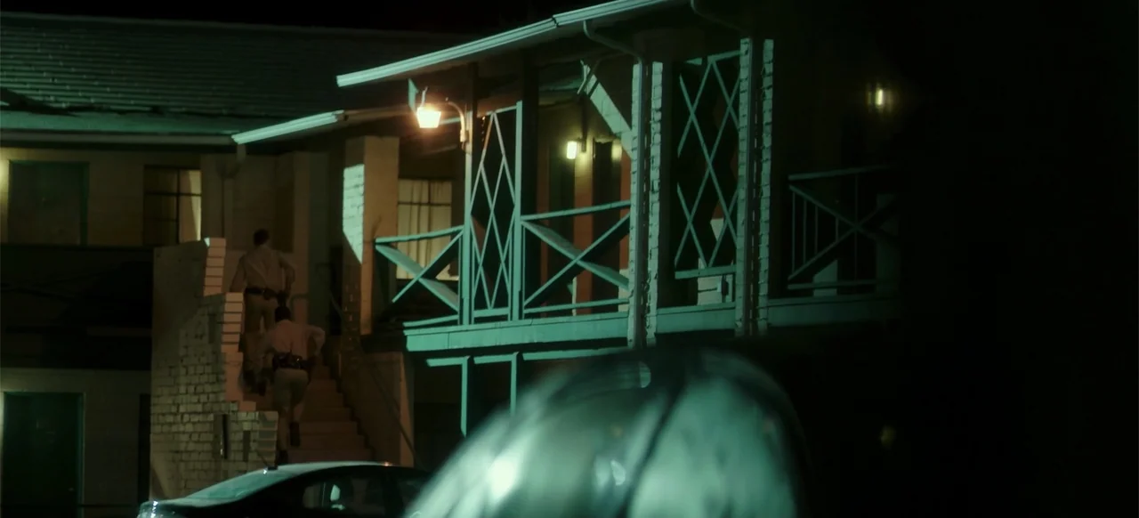 Blood Father (2016) movie still: wide — A multi-story motel building at night features wood-railed walkways and exterior staircases. Two uniformed…