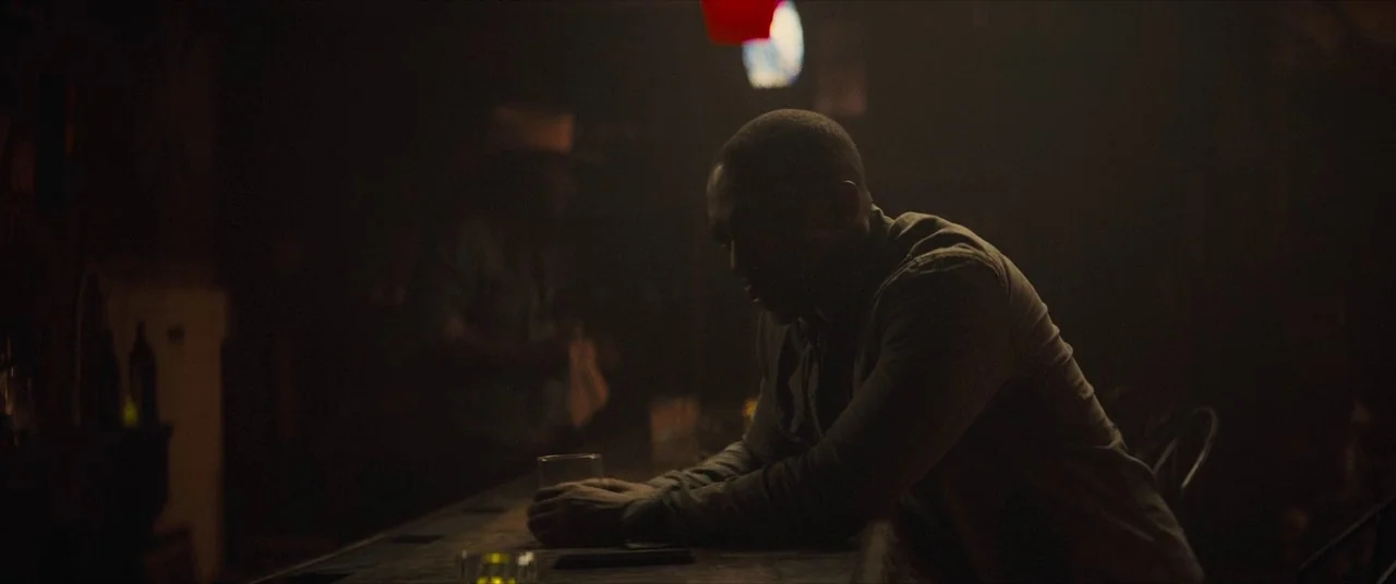 Synchronic (2019) movie still: medium — A dark dimly lit bar interior featuring a wooden counter and shadowy background figures. A solitary man…