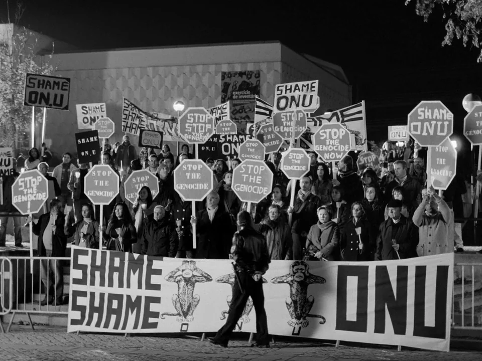 Tabu (2012) movie still: wide — A crowd of protestors holds numerous signs at night in a wide public square. A police officer in a leather jacket…