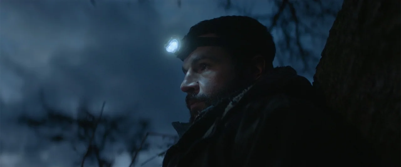 Bring Them Down (2025) movie still: closeup — A dark, wooded area at night with a cloudy sky. A man with a lit headlamp looks sideways while leaning…