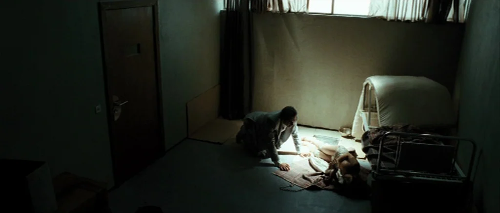 Adam Resurrected (2008) movie still: wide — A stark room features a heavy door and a low metal bed frame. A man kneels on the floor, reaching toward…