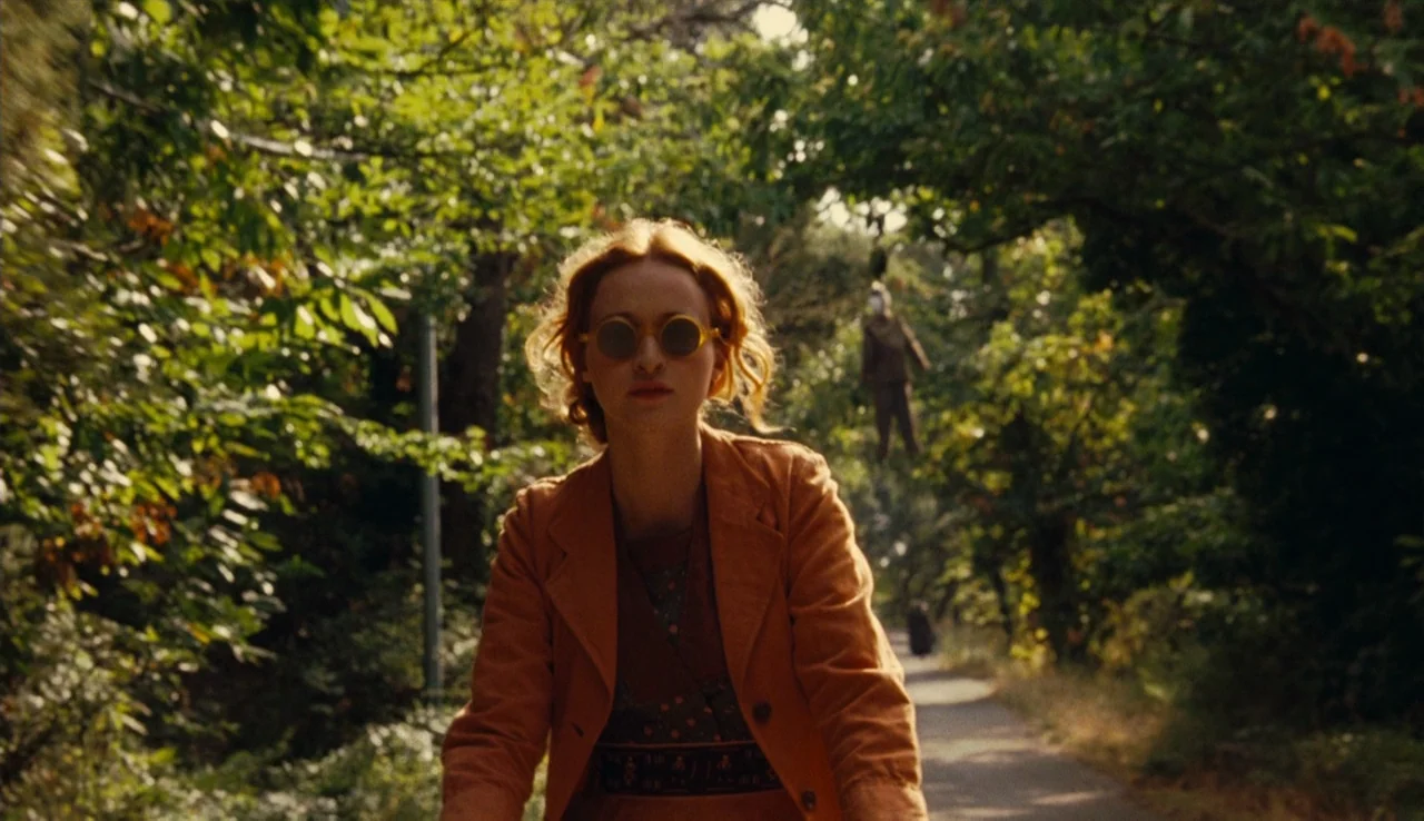 Renoir (2012) movie still: medium — A woman rides a bicycle along a path bordered by dense trees and greenery. An effigy hangs from a tree branch in…