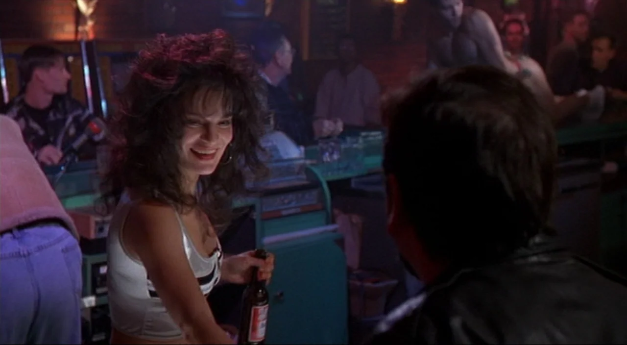 Pecker movie still: medium — A dimly lit 1990s establishment with brick walls and blurred figures in the background. A woman with voluminous hair…