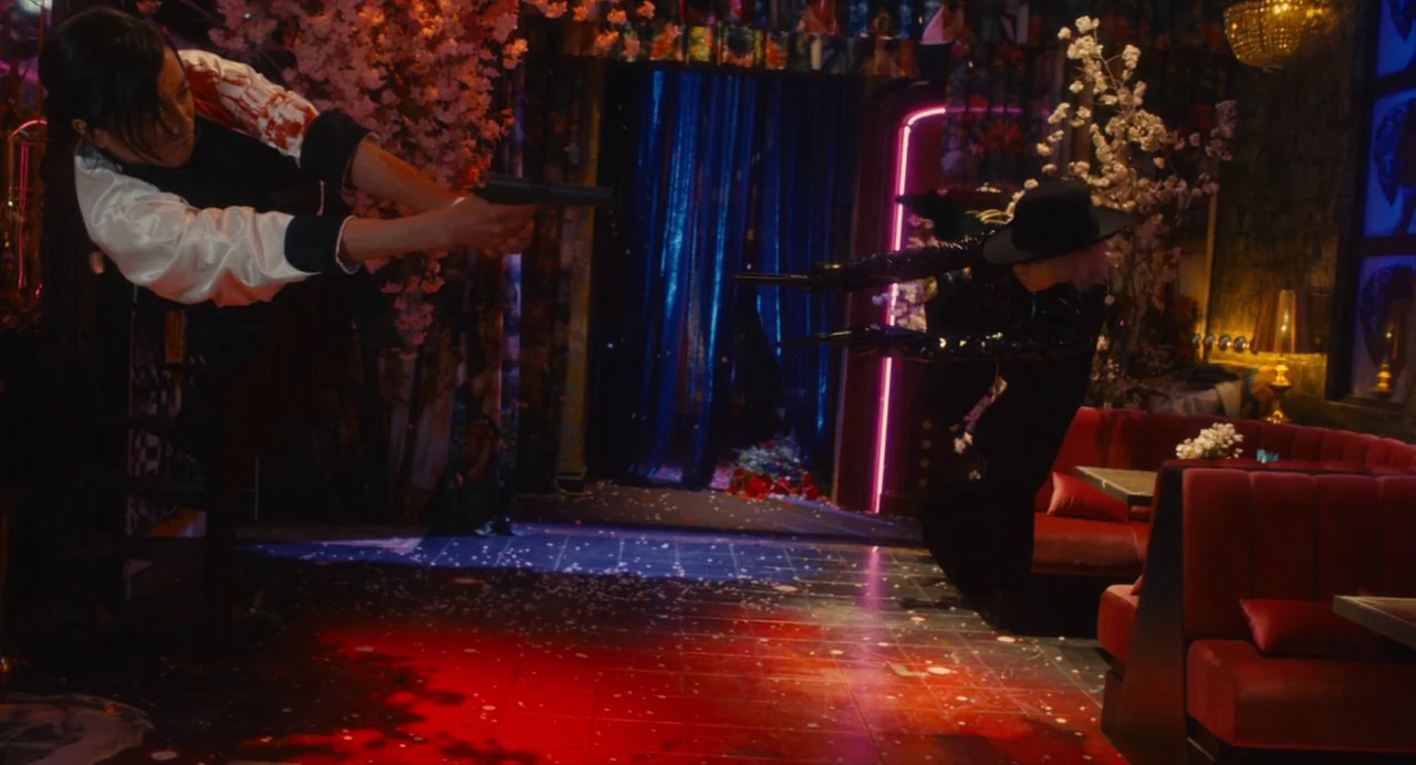 Diner (2019) movie still: wide — A dimly lit lounge decorated with pink cherry blossoms and velvet red furniture. Two individuals jump toward each…