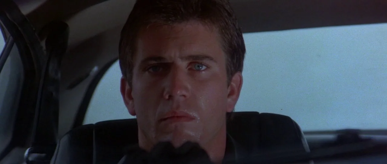 Mad Max (1979) movie still: closeup — A dark car interior shows a man sitting in the driver seat. He gazes straight forward with a faint sheen of…