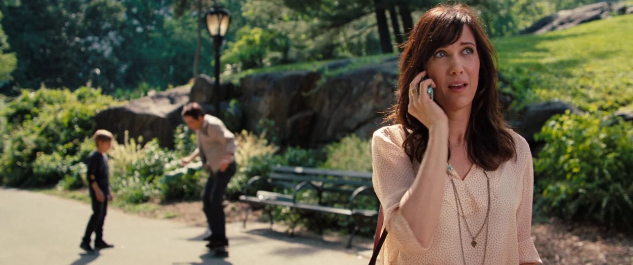 The Secret Life of Walter Mitty (2013) movie still: medium — A woman stands on a paved path in a lush green park. In the background a child and…