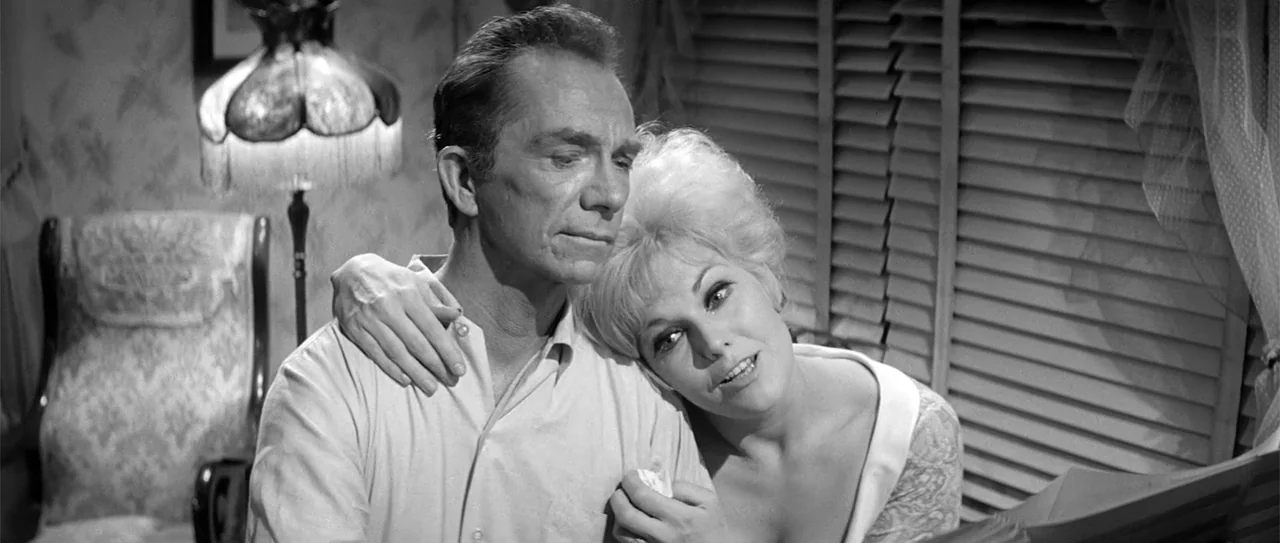 Kiss Me, Stupid (1964) movie still: medium — A domestic interior with a vintage floor lamp and window blinds in the background. A man sits while a…