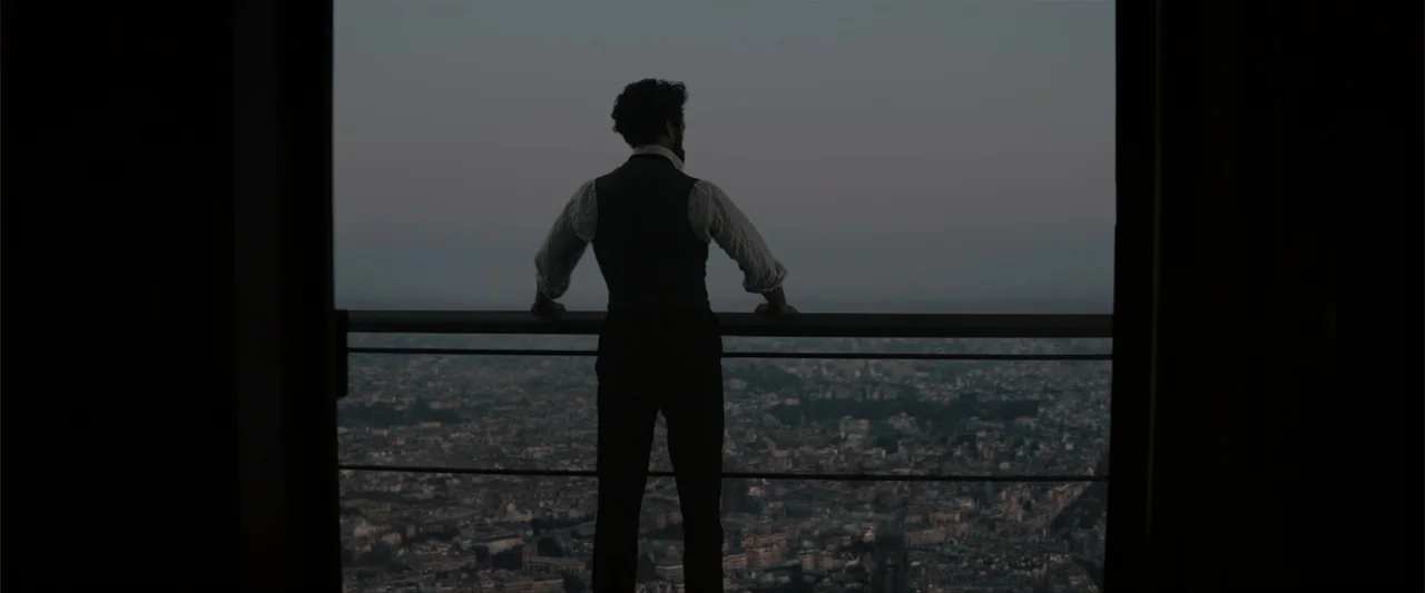 Eiffel (2021) movie still: fullbody — A man stands on a balcony overlooking a sprawling city at twilight. The dark silhouette of the figure frames…