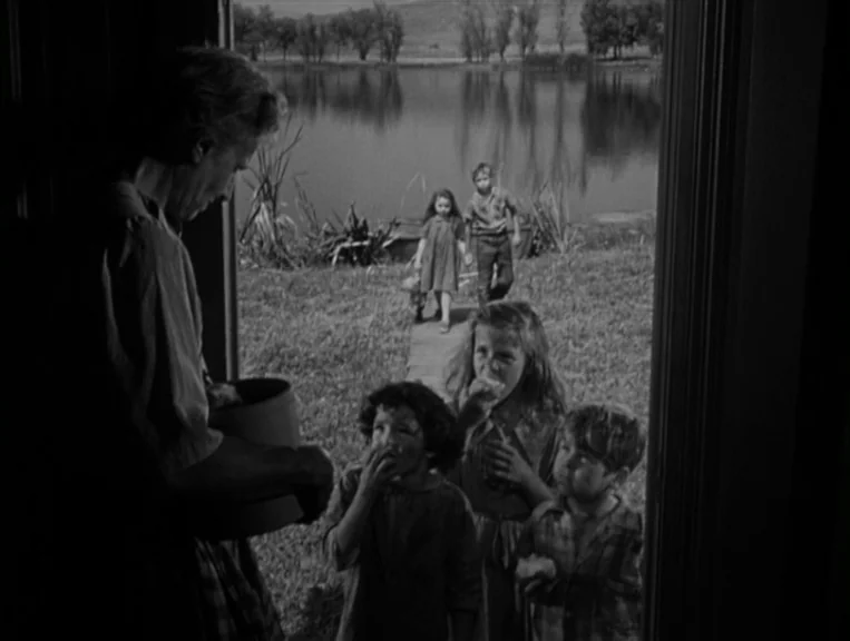 The Night Of The Hunter (1955) movie still: medium — A rural cabin doorway frames a woman handing items to a group of children. The background shows…