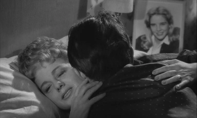 Lolita (1962) movie still: closeup — A dimly lit bedroom features a bed with pillows and a framed portrait photograph. A man and a woman are shown…