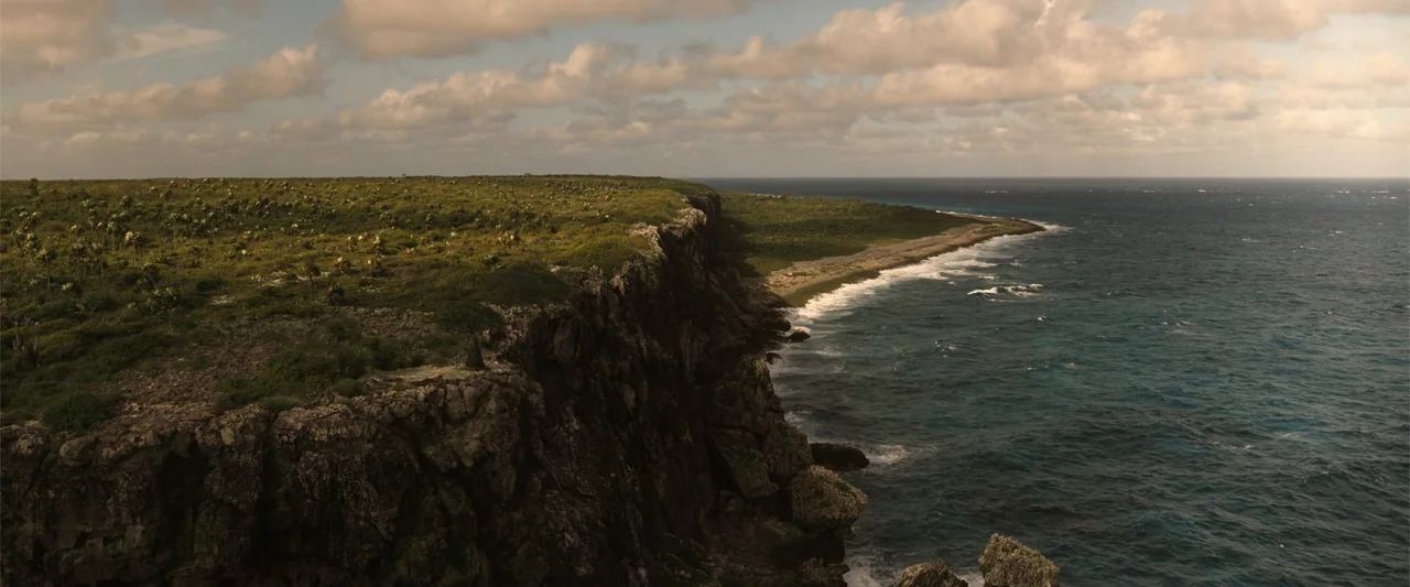 The Bluff (2026) movie still: establishing — A rugged limestone cliff overlooks a dark blue ocean under a cloudy sky. The coastline stretches into…
