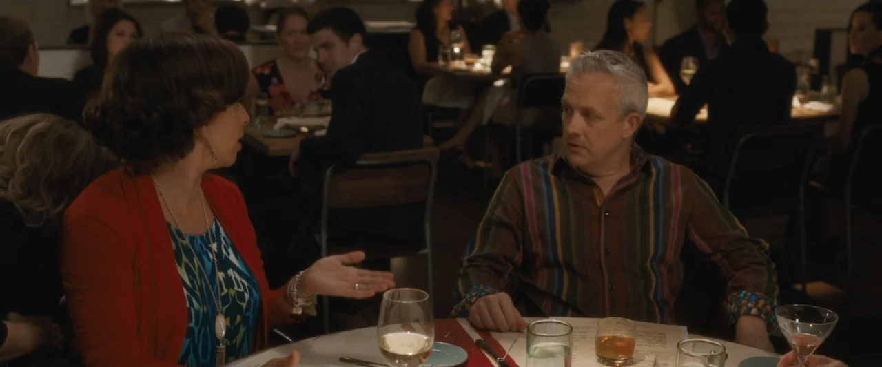 Life of the Party (2018) movie still: medium — A crowded restaurant with blurred diners in the background and a set table. A woman in a red cardigan…