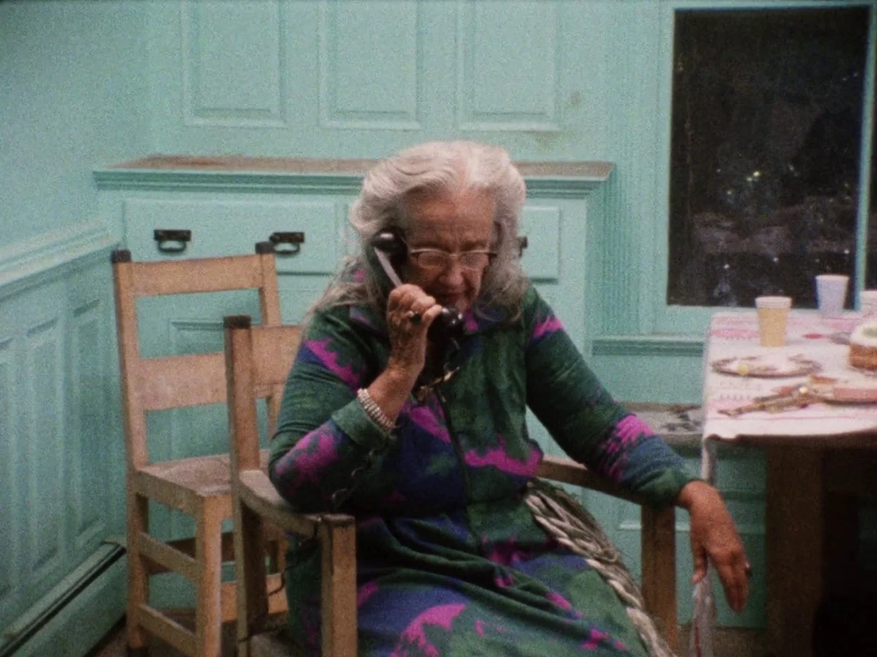 Grey Gardens (1975) movie still: medium — A room with mint-green paneled walls and a wooden chair sits behind the subject. The woman sits at a table…