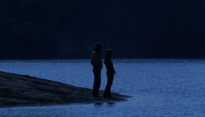 Let’s Scare Jessica To Death (1971) movie still: wide — A man and woman stand on a dark, sandy shoreline bordering a lake. They are silhouettes…