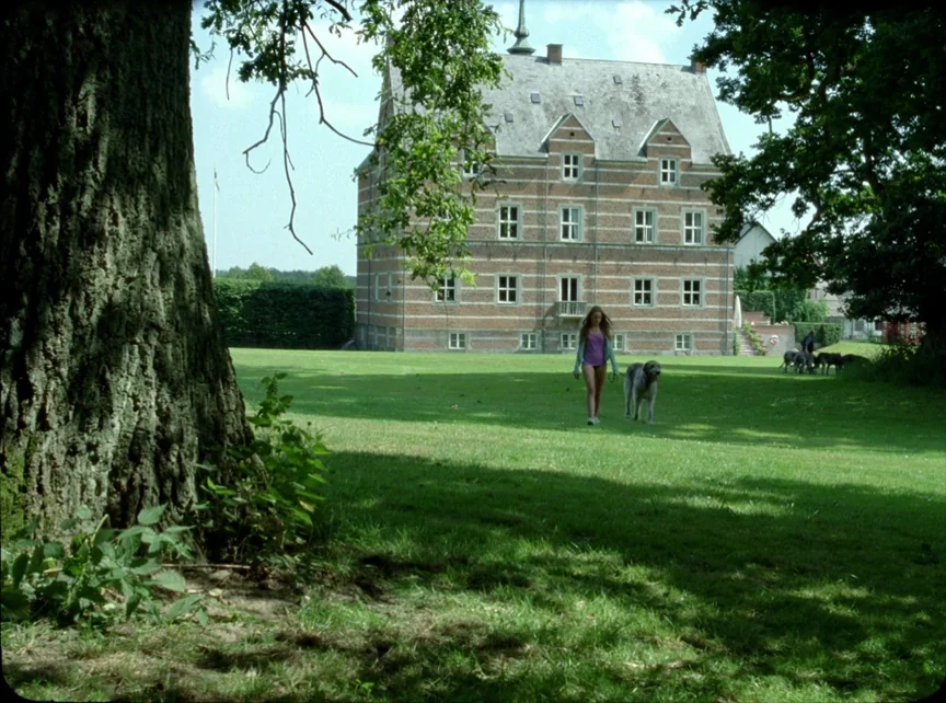 Jauja (2014) movie still: wide — A sprawling green lawn stretches before a classic brick mansion with multiple gabled windows. A young girl walks…