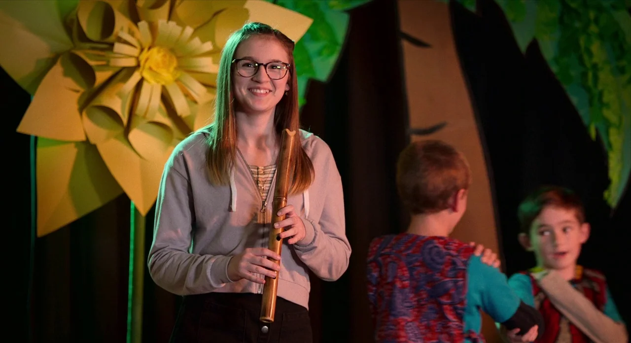 Where’d You Go Bernadette (2019) movie still: medium — A wooden stage features large paper flower props and dark fabric backdrops. A young girl in a…