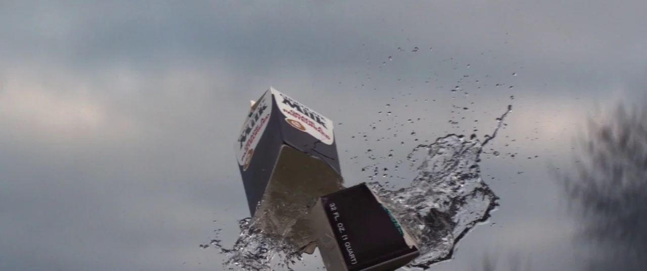 Super Dark Times (2017) movie still: closeup — A milk carton is captured mid-explosion against a muted, cloudy sky background. Water sprays outward…