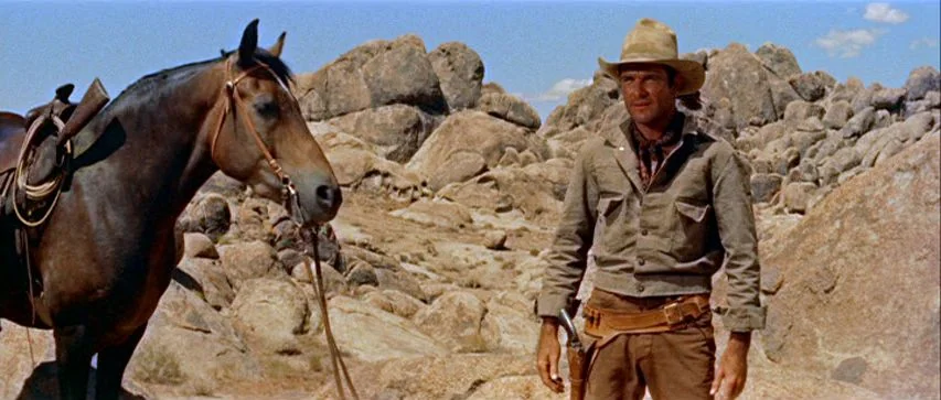 Ride Lonesome (1959) movie still: medium — A man and a horse stand in a barren desert landscape covered with boulders. The sunlit terrain is vast…