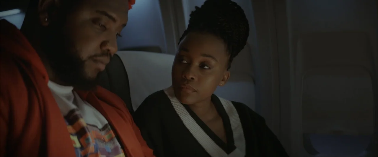 A Scam Called Love (2025) movie still: medium — The interior of an airplane cabin is dark with narrow seating visible. A man in a red hoodie and a…