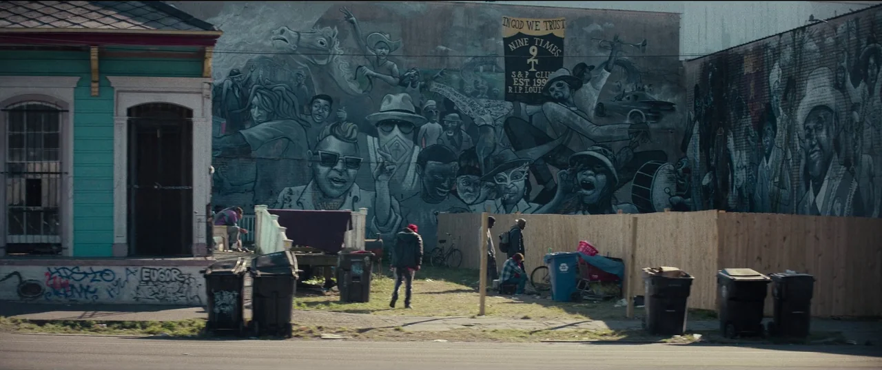 Hit Man (2023) movie still: wide — An urban lot features a large painted mural on the exterior wall of a building. Several people gather near a…