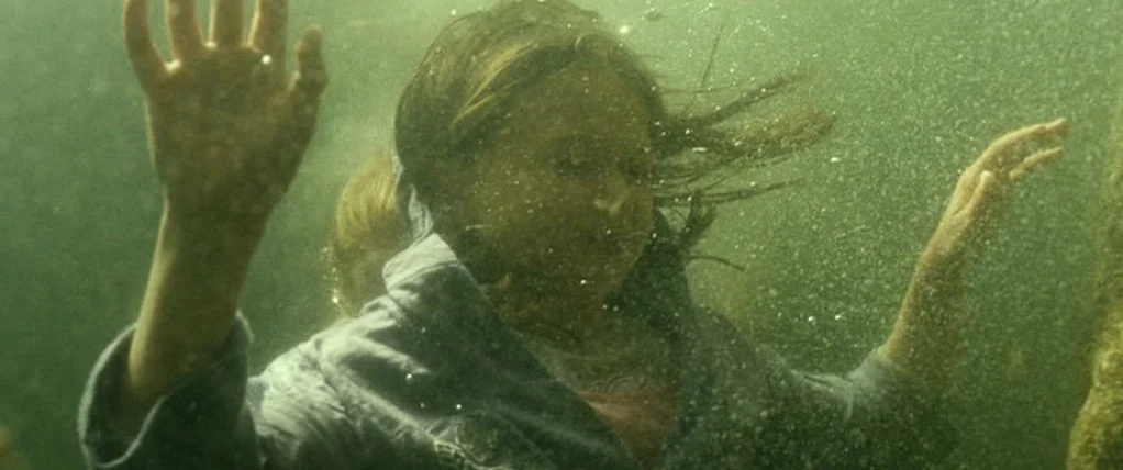 Ondine (2009) movie still: medium — A young girl is submerged beneath the surface of murky, green-tinted lake water. Her hands are extended upward…