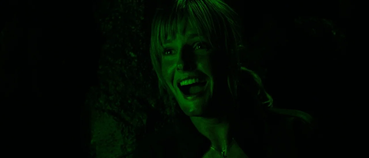 The Descent (2005) movie still: closeup — A woman stands in a dark cave environment with rough, shadowed stone walls. She has her mouth wide open…