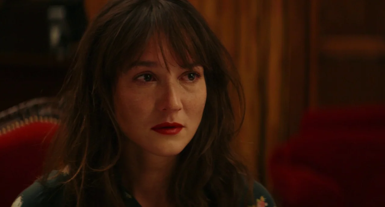 Anaïs in Love (2021) movie still: closeup — A dimly lit room with wood-paneled walls and a red upholstered chair. A woman with long brown hair looks…