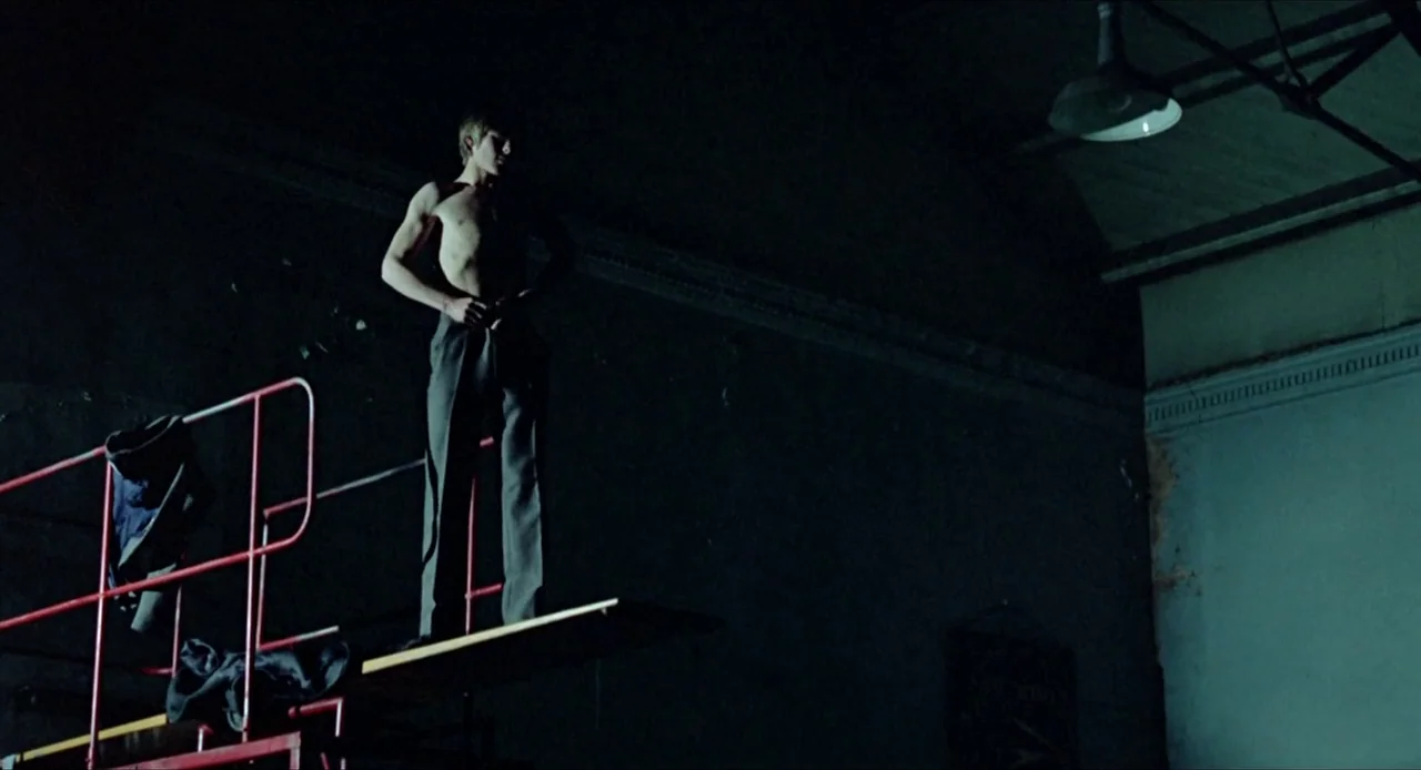 Deep End (1970) movie still: fullbody — A dark indoor swimming facility features a metal diving board structure against high walls. A shirtless man…